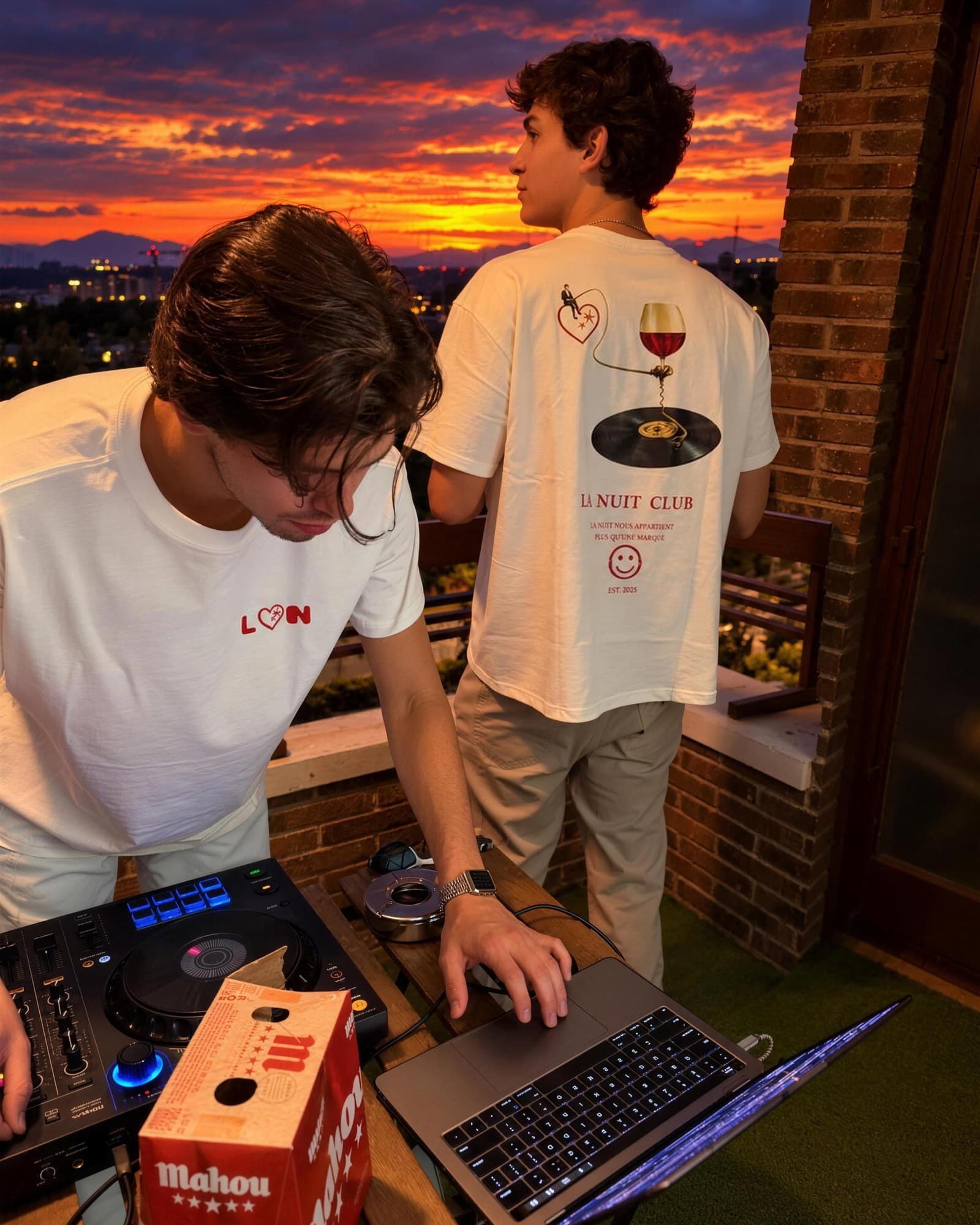 Dos chicos con la camiseta La Nuit en un balcón al atardecer; uno pincha música.
