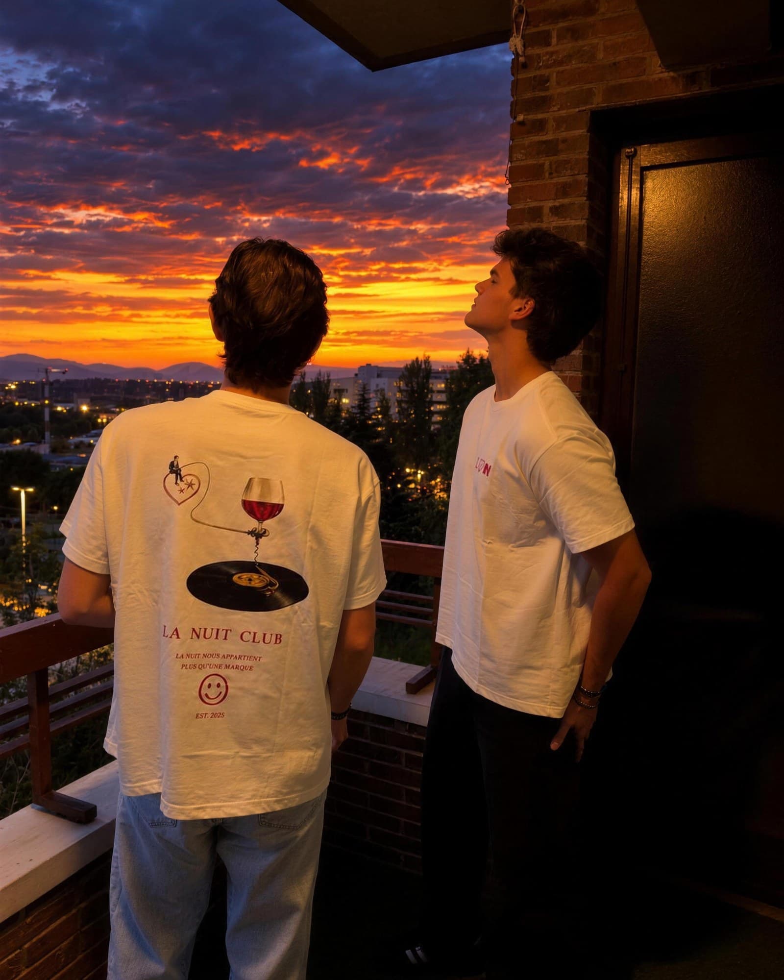 Dos chicos con la camiseta La Nuit miran el atardecer desde un balcón.
