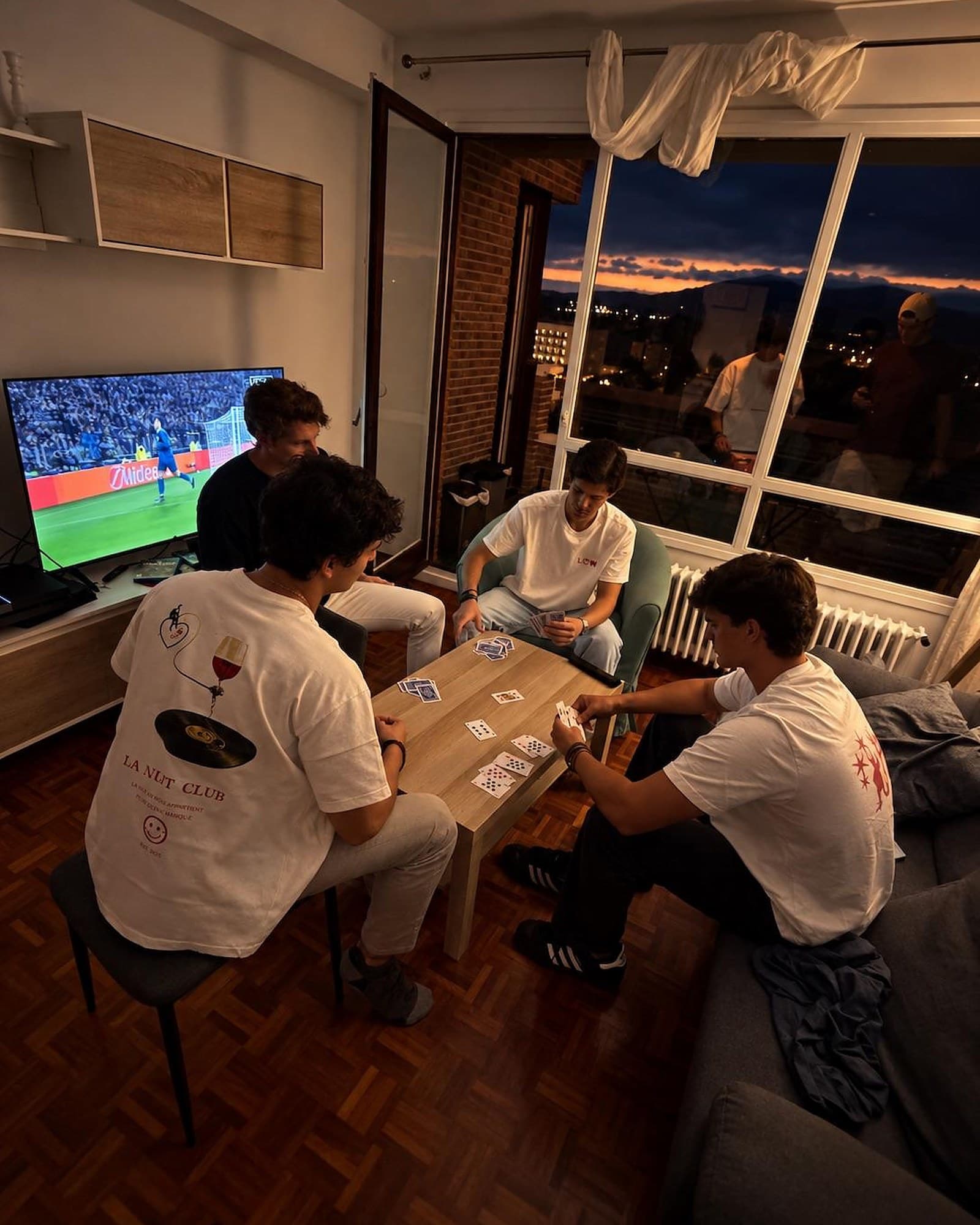 Cuatro amigos juegan a las cartas al atardecer; uno lleva la camiseta La Nuit con el cartel del club en la espalda.