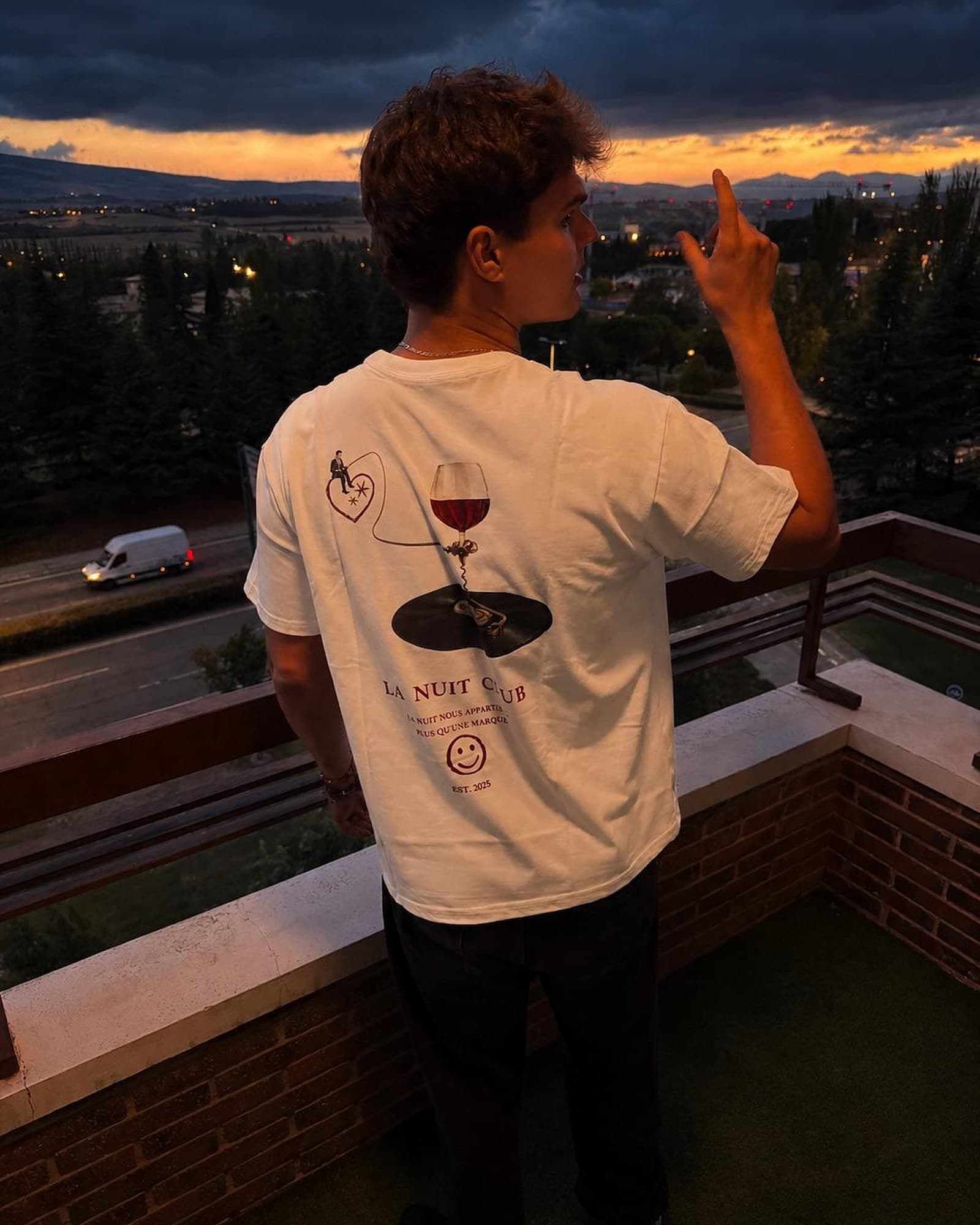 Un chico de espaldas en un balcón, con el cartel del club de la camiseta La Nuit bien visible, señala el atardecer sobre la ciudad.