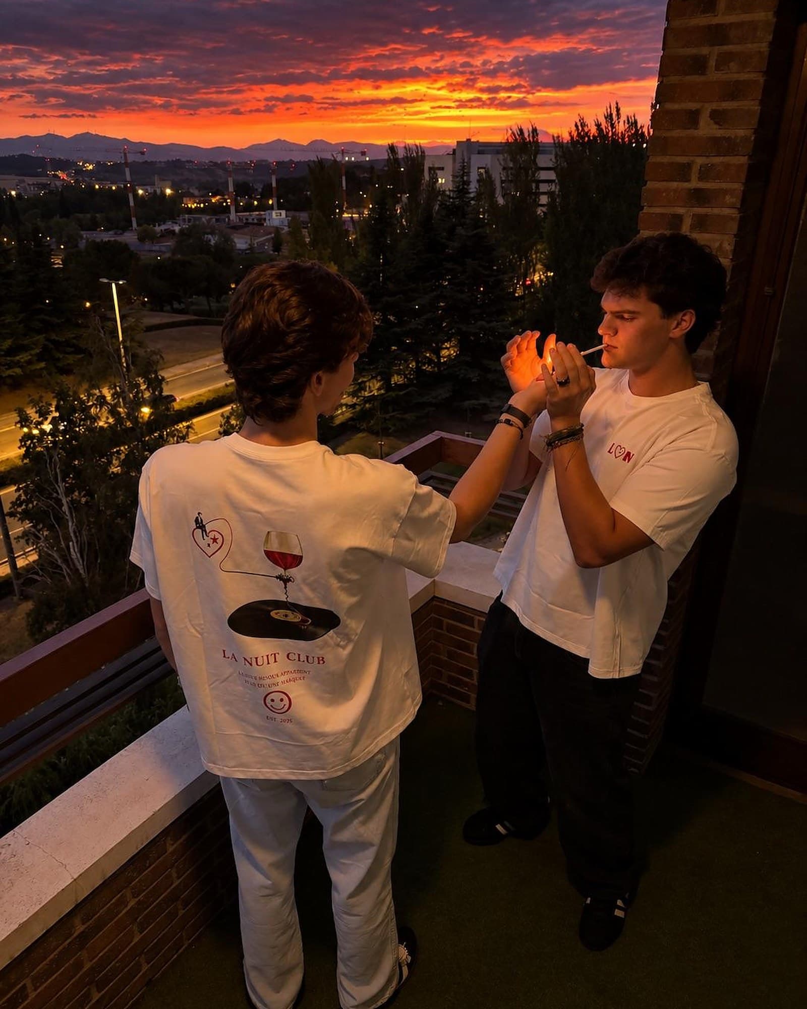 Dos chicos en un balcón al atardecer: uno enseña el cartel del club en la espalda de la camiseta La Nuit y el otro lleva el logo en el pecho.