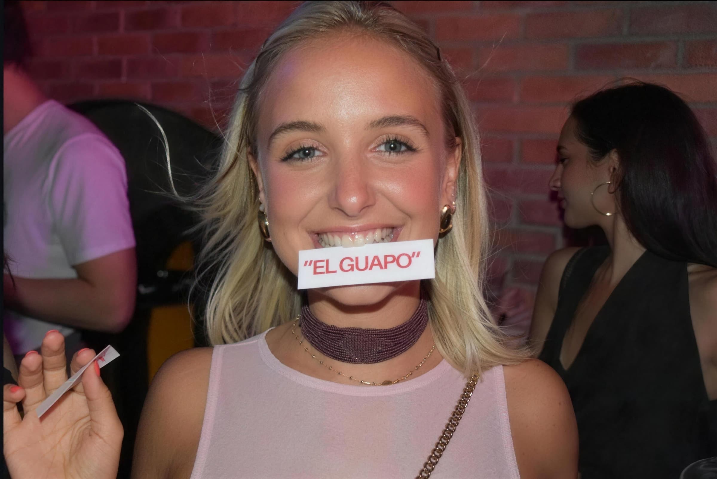 Una chica sonríe en una fiesta con una pegatina que dice «El Guapo» entre los dientes.