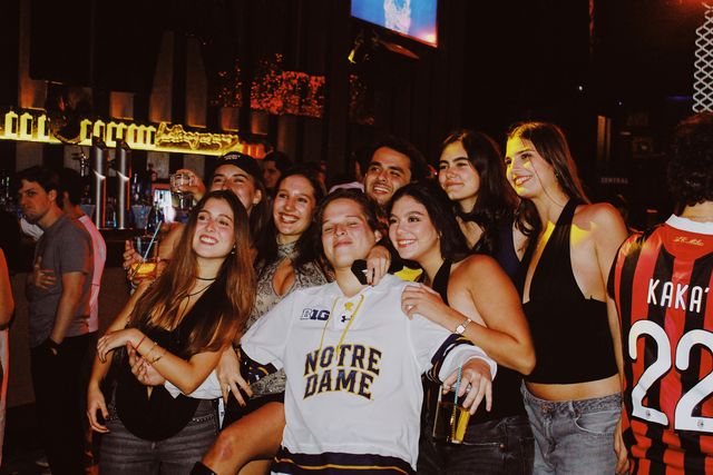 Chicos y chicas posan apiñados y sonríen junto a la barra iluminada; en el centro, alguien con camiseta blanca de hockey, y a la derecha, un chico de espaldas.