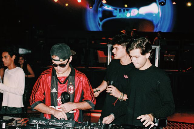 El DJ de camiseta de fútbol a rayas rojas y negras, con gafas de sol, sostiene un auricular de mano junto a dos chicos de negro, bajo una pantalla azul.