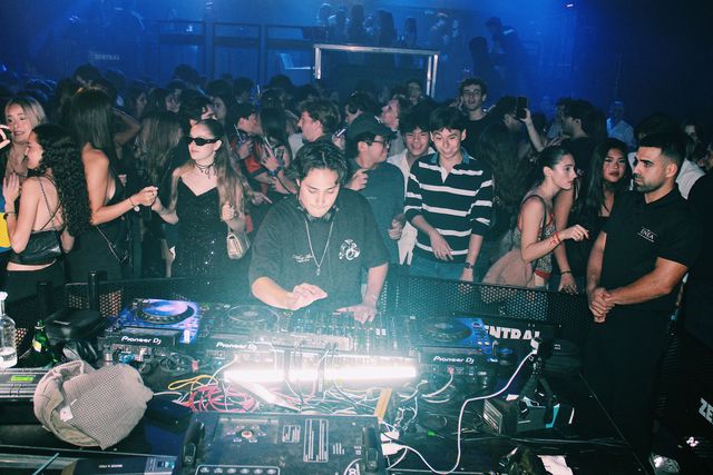 Un DJ con camiseta negra y cadena al cuello mezcla con la vista en la mesa, rodeado de cerca por el público bajo luz azul.