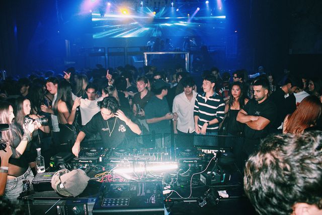 Un DJ con camiseta negra y cadena al cuello se lleva una mano a la cabeza mientras mezcla, con el público alrededor y una pantalla LED azul al fondo.