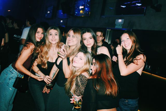 Siete chicas posan muy juntas ante una pared oscura con pantallas; la mayoría sonríe a cámara, una levanta un vaso y la del top de leopardo pone morritos.