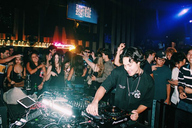 Un DJ con camiseta negra y cadena al cuello sonríe mientras mezcla, con el público pegado a la cabina y una pantalla con el nombre de Zentral al fondo.