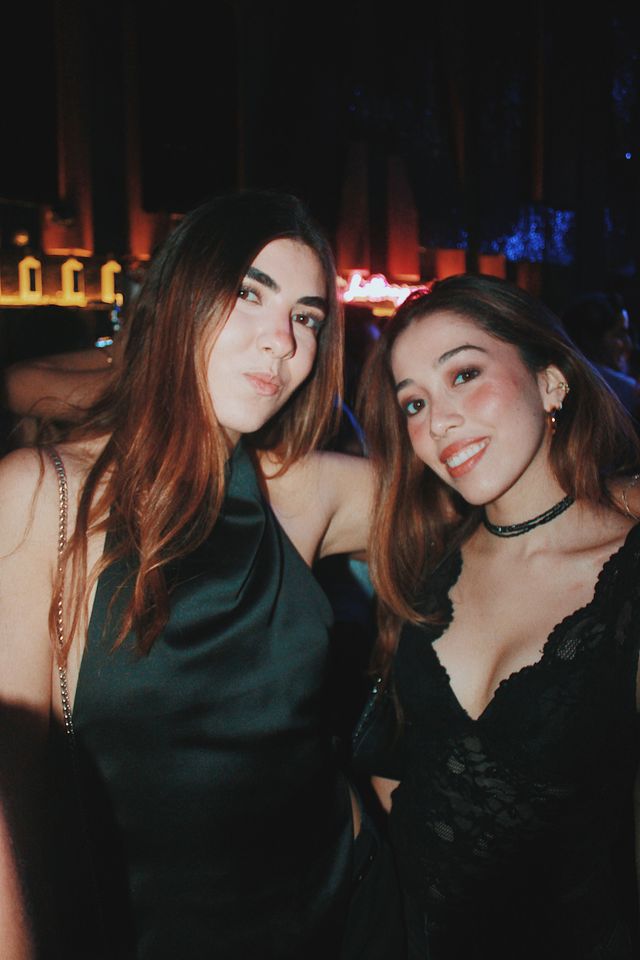 Dos chicas posan muy juntas; la del top negro satinado pone morritos y la del top de encaje negro con gargantilla sonríe a cámara, con luces cálidas al fondo.