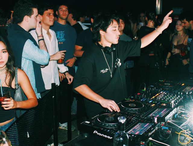 Un DJ con camiseta negra y cadena al cuello levanta un brazo y señala hacia arriba mientras mezcla, con el público tras la valla de la cabina.