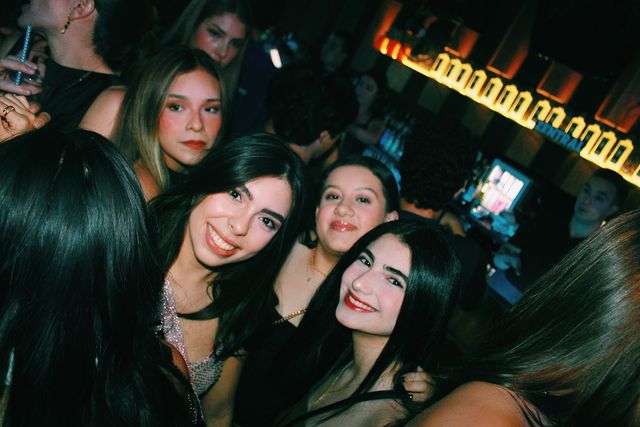 Varias chicas posan apretadas junto a la barra iluminada; tres sonríen a cámara, una con top de lentejuelas, y detrás otra de melena clara mira seria.
