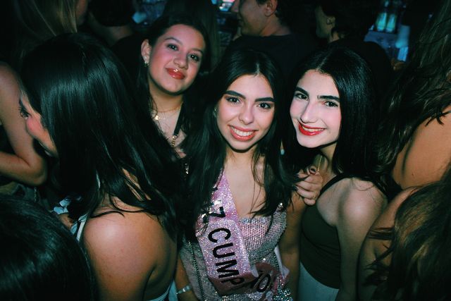 Tres chicas sonríen a cámara entre la multitud; la del centro lleva un top rosa de lentejuelas con una banda de cumpleaños, y a la izquierda otra chica aparece de perfil.