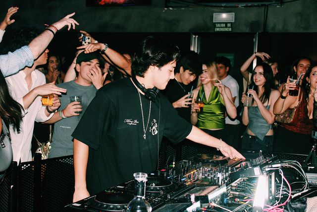 Un DJ con camiseta negra y cadena al cuello mezcla mientras el público levanta brazos y vasos tras la valla; a la derecha ríe una chica de top verde.