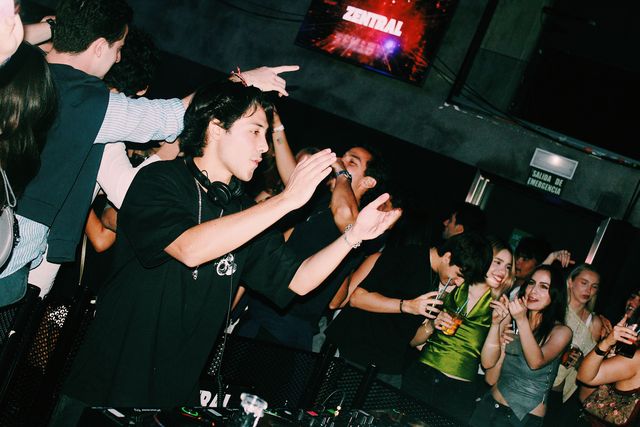 Un DJ con camiseta negra y cadena al cuello extiende las manos hacia el público, con la pantalla de Zentral al fondo; a la derecha ríe una chica de top verde.