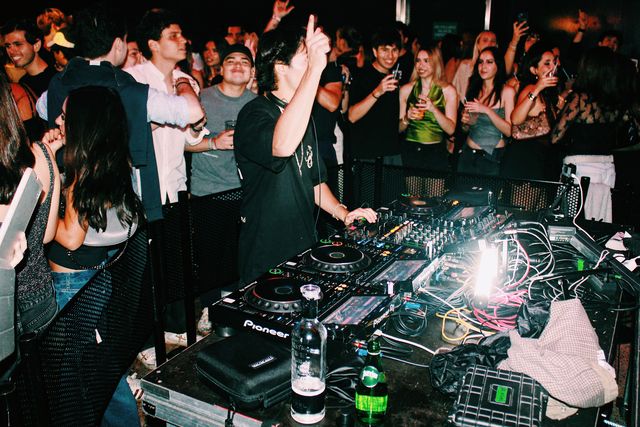 Un DJ con camiseta negra y cadena al cuello señala hacia arriba mientras mezcla, con botellas sobre la mesa en primer plano y el público levantando vasos y móviles detrás.