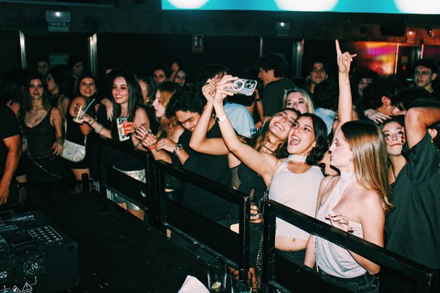 Tras la valla de la cabina, dos chicas se hacen un selfi entre risas y otra, de perfil, levanta el índice, rodeadas de público con vasos en la mano.