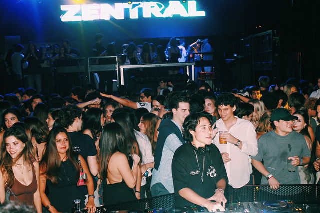 Un DJ con camiseta negra y cadena al cuello mezcla rodeado por la multitud bajo el gran letrero luminoso de Zentral, con más público en una plataforma al fondo.