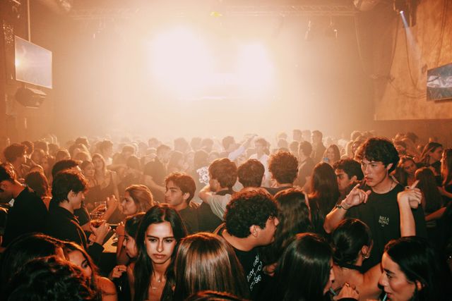 La multitud llena la pista entre humo y luz anaranjada; delante, una chica de melena oscura mira a cámara y un chico de negro señala con el dedo.