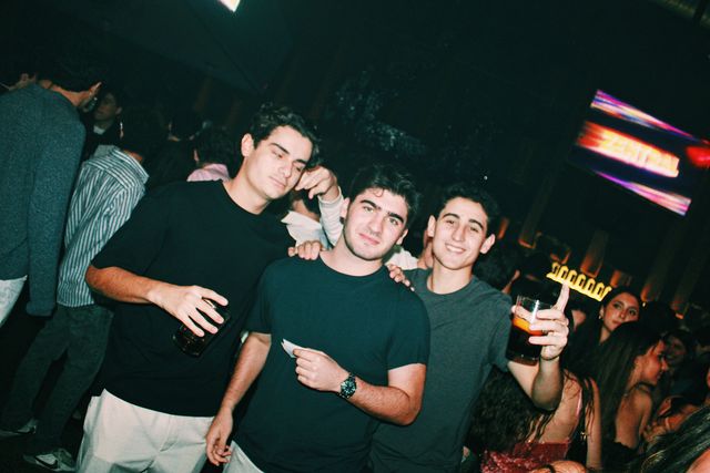 Tres chicos posan con la pantalla de Zentral al fondo; el de camiseta gris sonríe y levanta un vaso, y el de la izquierda, con otro vaso, entorna los ojos.