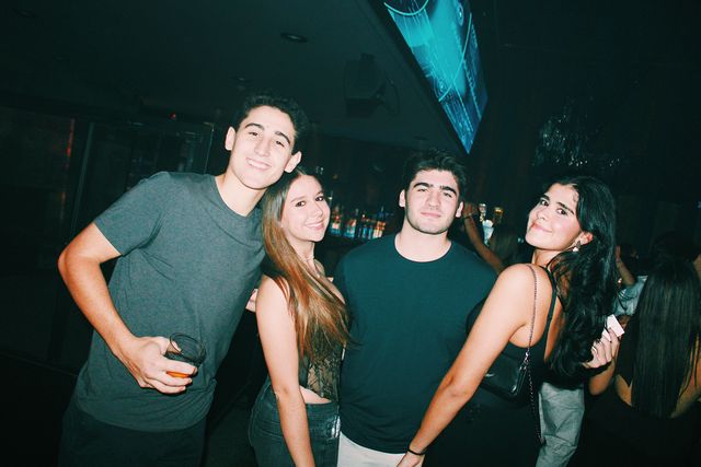 Dos chicos y dos chicas posan intercalados bajo una pantalla azul; el de camiseta gris sostiene un vaso y la chica del bolso negro mira a cámara por encima del hombro.