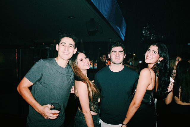 Dos chicos y dos chicas posan ante la barra iluminada; la de melena castaña ladea la cabeza hacia el chico de camiseta gris y la del bolso negro sonríe a cámara.