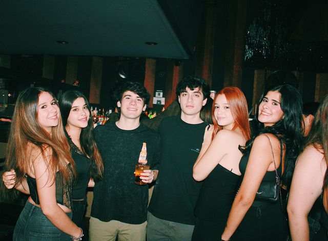 Cuatro chicas y dos chicos posan junto a la barra y casi todos sonríen a cámara; el chico de pantalón beige sostiene una botella y un vaso.