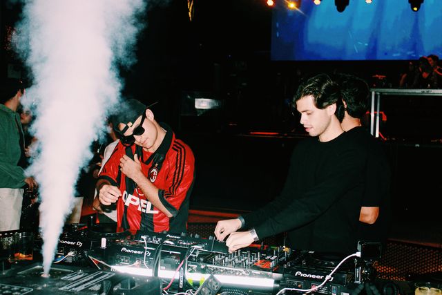 El DJ de camiseta de fútbol a rayas rojas y negras escucha con un auricular de mano y un chico de jersey negro mezcla junto a un chorro de humo.