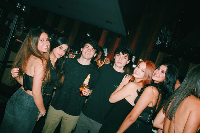 Cuatro chicas y dos chicos posan apiñados junto a la barra en un encuadre inclinado; casi todos sonríen, y la pelirroja y la del bolso negro acercan las cabezas.