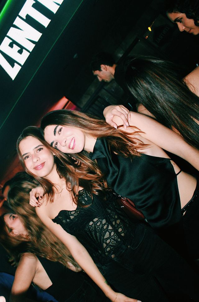 Dos chicas posan juntas y sonríen a cámara bajo el letrero luminoso de Zentral; una lleva un top de encaje negro y la otra, un top negro satinado.