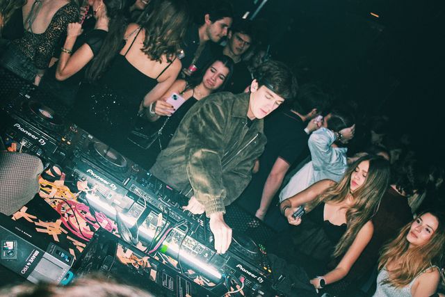 Un DJ con chaqueta verde oliva mezcla con la mirada baja, rodeado de cerca por chicos y chicas; una de ellas sostiene un móvil rosa junto a los platos.