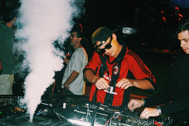 De cerca, el DJ de camiseta de fútbol a rayas rojas y negras, con gafas de sol, baja la vista junto a un chorro de humo blanco.
