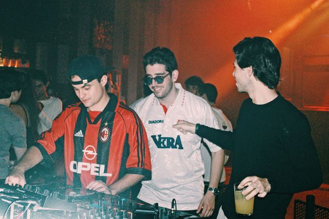 El DJ de camiseta de fútbol a rayas rojas y negras mezcla y un chico de jersey negro señala a otro con gafas de sol y camiseta blanca, bajo luz naranja.