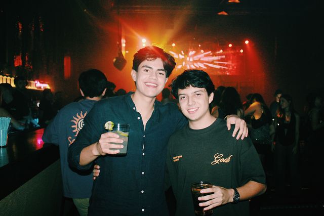 Dos chicos, uno con camisa oscura y otro con camiseta, posan abrazados y sonríen a cámara, vaso en mano, ante una pantalla y luces rojas.