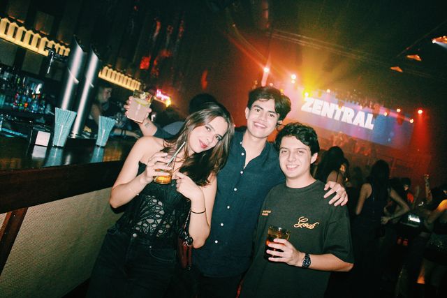 Una chica con top negro de encaje y dos chicos sonríen a cámara junto a la barra, con un vaso en alto y Zentral en la pantalla del fondo.