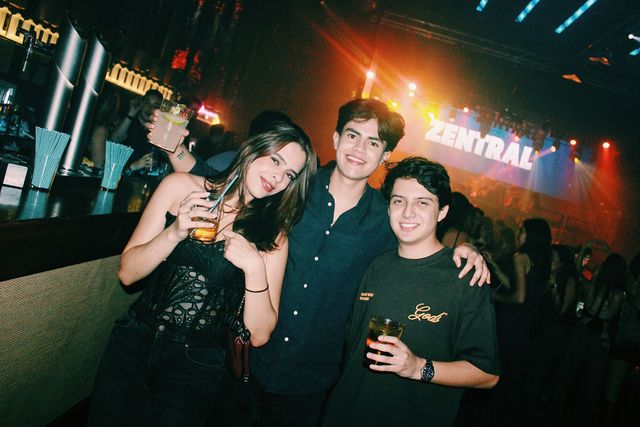 Una chica con top negro de encaje ladea la cabeza y sonríe junto a dos chicos sonrientes en la barra, con un vaso alzado y Zentral en la pantalla.