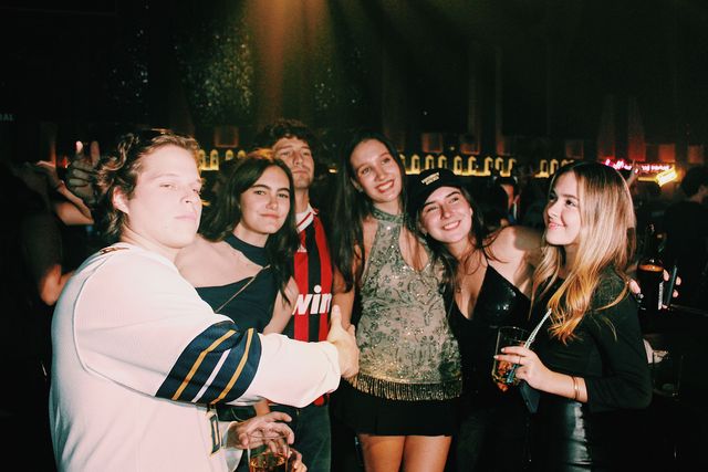 Cuatro chicas y dos chicos posan con la barra iluminada al fondo; tres chicas sonríen a cámara y un chico con camiseta blanca de hockey extiende el brazo hacia ellas.