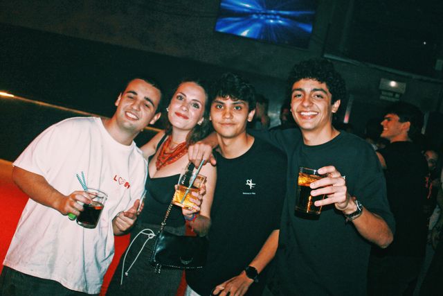 Tres chicos y una chica con collar rojo posan con sus bebidas bajo una pantalla azul, la mayoría sonriendo a cámara.