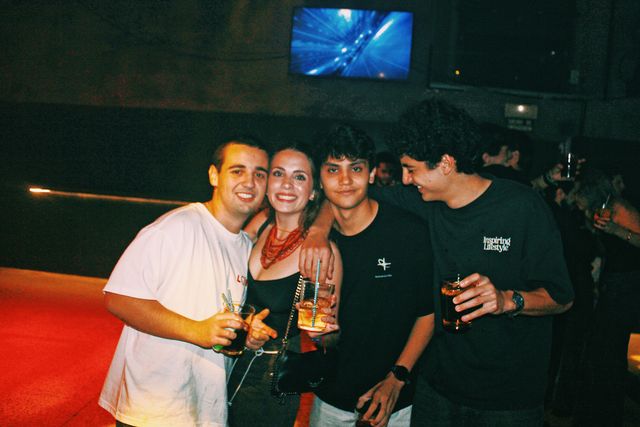 Tres chicos y una chica con collar rojo posan; ella y el de camiseta blanca sonríen a cámara y el de pelo rizado se ríe mirando hacia abajo.