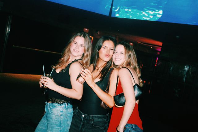 En plano inclinado, tres chicas posan muy juntas bajo una pantalla azul; la del centro pone morritos y las otras dos sonríen a cámara.