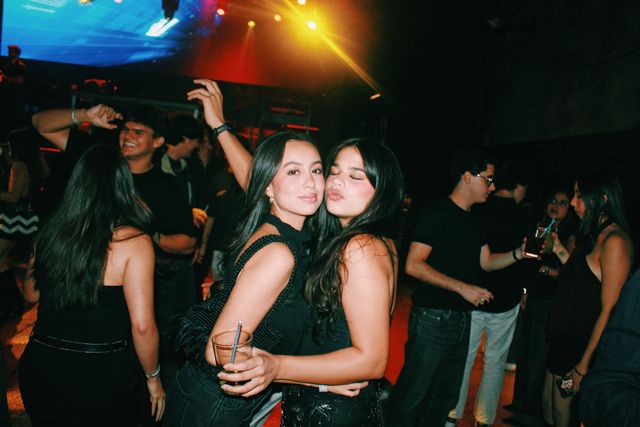 Dos chicas de negro posan mejilla con mejilla entre el público, una mirando a cámara y la otra con los ojos cerrados y morritos, bajo luces de colores.