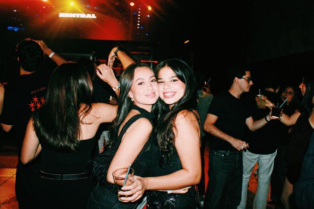 Dos chicas de negro posan mejilla con mejilla y sonríen a cámara, con un vaso en la mano y el letrero de Zentral entre luces rojas al fondo.