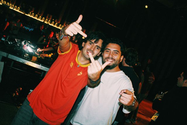 En plano inclinado, dos chicos posan junto a la cabina: uno con camiseta roja de cuello amarillo hace la peineta con las dos manos y el otro, de camiseta blanca, sonríe.