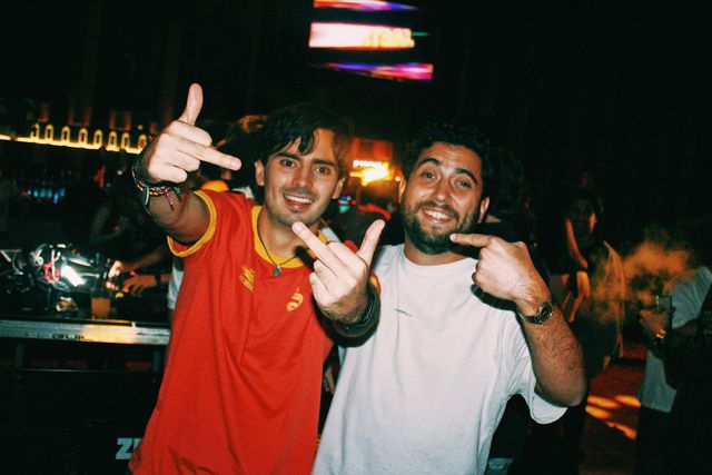 Dos chicos sonríen a cámara junto a la cabina: uno con camiseta roja de cuello amarillo hace la peineta con las dos manos y el de camiseta blanca señala con el dedo.