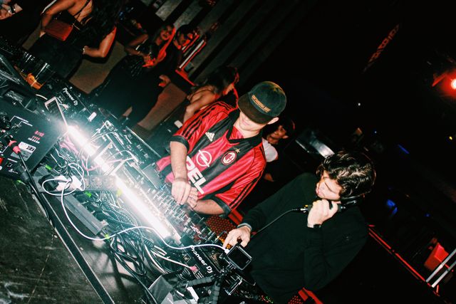 En plano inclinado, el DJ de camiseta de fútbol a rayas rojas y negras mezcla y un chico de jersey negro se lleva un auricular de mano al oído.