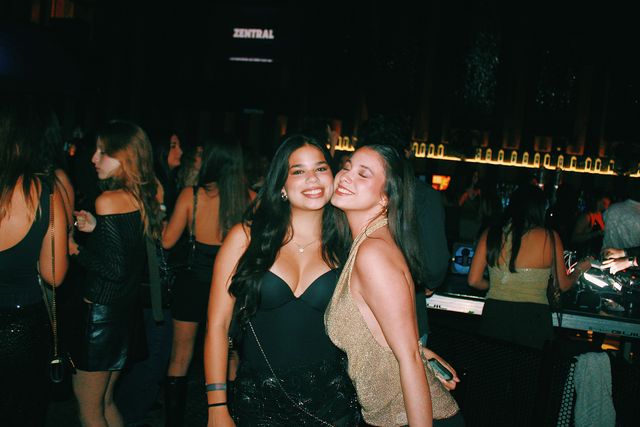 Dos chicas juntan las mejillas: la de negro sonríe a cámara y la de top dorado sonríe con los ojos cerrados; al fondo, la barra y el letrero de Zentral.
