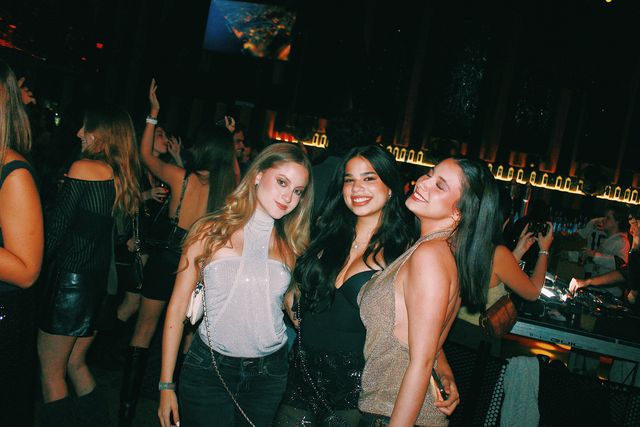 Tres chicas posan junto a la barra iluminada: la de top blanco brillante mira a cámara, la de negro sonríe y la de top dorado sonríe con los ojos cerrados.