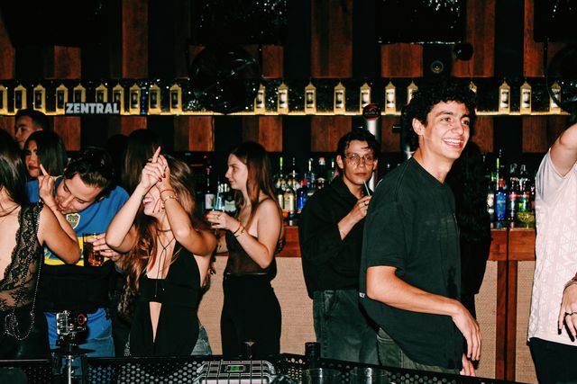 Delante de la barra, con botellas iluminadas y el letrero de Zentral, un chico de camiseta negra sonríe y una chica se ríe tapándose los ojos, entre más gente con vasos.