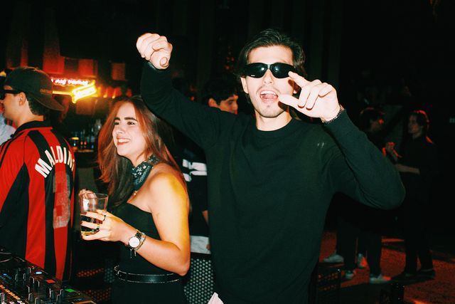 Un chico de negro con gafas de sol alza el puño junto a una chica sonriente con pañuelo al cuello y al DJ de camiseta de fútbol a rayas rojas y negras.