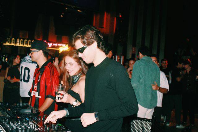Un chico de negro con gafas de sol mezcla en los platos junto a una chica sonriente con un vaso y al DJ de camiseta de fútbol a rayas rojas y negras.