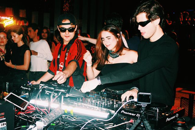 Tras los cables y un tubo de luz blanca, el DJ de camiseta de fútbol a rayas rojas y negras y un chico de negro mezclan, con una chica bailando entre ambos.